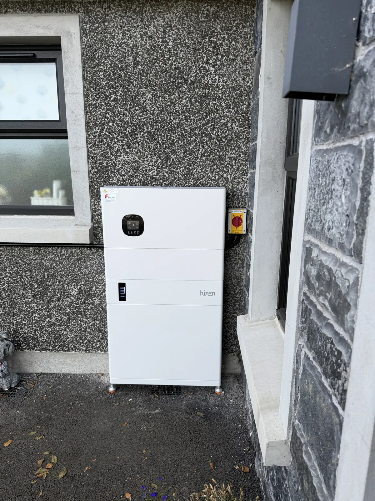 Hinen all-in-one battery storage unit installed outdoors against a pebble-dash wall