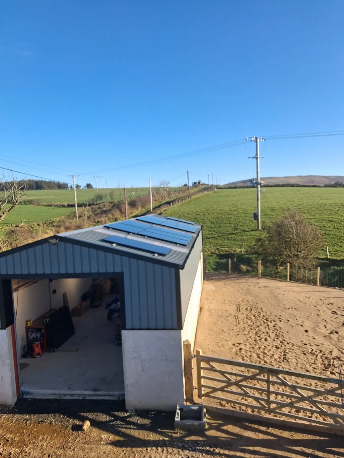Agricultural solar panels on the roof of a farm outbuilding beside a sandy yard, with fields behind